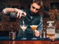 Barman przelewa drinka do kieliszka, pokazując, jak być dobrym barmanem. Na blacie stoją też inne drinki i akcesoria.