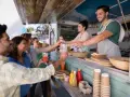 Uśmiechnięci klienci odbierają napoje od barmana w food trucku. Ciekawe, ile zarabia barman w takim miejscu?