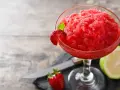 Orzeźwiający strawberry daiquiri, udekorowany plasterkiem truskawki i listkiem mięty, obok połówki limonki i świeżej truskawki.