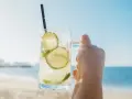 Orzeźwiająca Caipirinha czy Caipiroska z limonką i lodem, trzymana na tle plaży i błękitnego nieba. Idealny napój na wakacje.