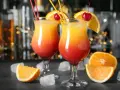Dwa kolorowe drinki z parasolką, ozdobione pomarańczą i wiśnią, stoją na barze.