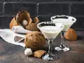 Dwa kieliszki drinka z malibu z mlekiem, ozdobione wiórkami kokosowymi, obok świeżych kokosów.
