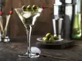Elegancki drink z martini bianco, ozdobiony trzema oliwkami na patyczku. Obok kostki lodu i talerzyk z oliwkami.