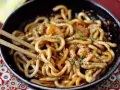 Miseczka z makaronem udon z krewetkami, posypana sezamem i pietruszką. Pałeczki zanurzone w daniu. Prosty przepis na to, jak gotować makaron udon.