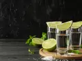 Trzy kieliszki z drinkiem z mezcalem lub tequilą, ozdobione ćwiartkami limonki i miętą, na tle czarnej ściany.