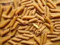 Makaron pełnoziarnisty vs zwykły. Na drewnianym tle leży mnóstwo makaronu penne. Wśród nich wyróżnia się jeden, żółty makaron fusilli.