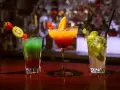 Trzy kolorowe drinki na barze. Jeden z nich to drink z pomarańczą, ozdobiony owocami.