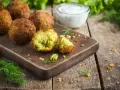 Chrupiące falafelki na desce, jeden przekrojony, ukazujący zielone wnętrze. Obok miseczka sosu i świeży koperek. Idealny falafel przepis.