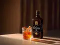 Whiskey z colą kontra Whiskey z ginger ale. Szklanka z bursztynowym trunkiem i butelka Ballantine's 10 Years Old.
