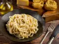 Pyszne Fettuccine Alfredo przepis na talerzu, obok surowy makaron, ser i oliwa. Idealne danie dla miłośników włoskiej kuchni.