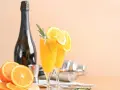 Dwa kieliszki z pomarańczowym napojem, ozdobione plasterkami pomarańczy i rozmarynem. Obok butelka szampana, shaker i świeże pomarańcze. Mimosa czy Bellini?