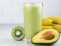 Zielony, kremowy koktajl z awokado, kiwi i banana. Obok szklanki połówka awokado i przekrojone kiwi. W tle pęk bananów.
