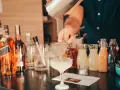 Barman przelewa płyn z shakera przez sitko do kieliszka, tworząc koktajl. W tle butelki alkoholi i kolorowe syropy.