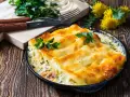 Pyszne conchiglioni zapieczone z serem i ziołami, podane w czarnej patelni. Obok deska z nożem i widelcem, oraz pęczek natki pietruszki.