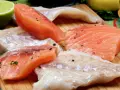 Stek z łososia kontra polędwica z dorsza. Surowe kawałki ryb, przyprawione pieprzem, leżą obok limonki, cytryny i ziół.