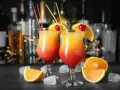 Dwa drinki z wódką, ozdobione pomarańczą i wiśnią, z kostkami lodu i owocami na blacie.
