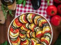 Kolorowy ratatouille przepis w naczyniu, ułożony z plastrów cukinii, bakłażana i pomidorów. Obok świeże warzywa i zioła.