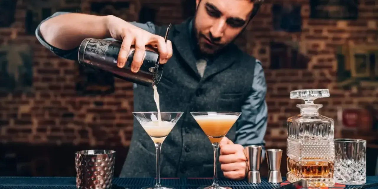 Barman przelewa drinka do kieliszka, pokazując, jak być dobrym barmanem. Na blacie stoją też inne drinki i akcesoria.