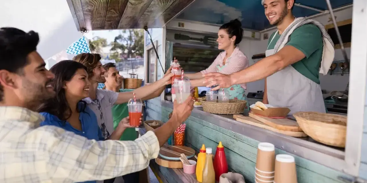 Uśmiechnięci klienci odbierają napoje od barmana w food trucku. Ciekawe, ile zarabia barman w takim miejscu?