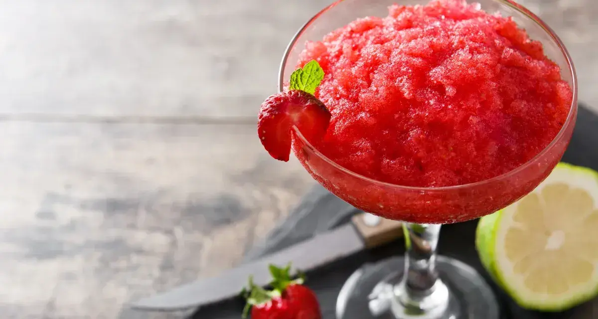 Orzeźwiający strawberry daiquiri, udekorowany plasterkiem truskawki i listkiem mięty, obok połówki limonki i świeżej truskawki.