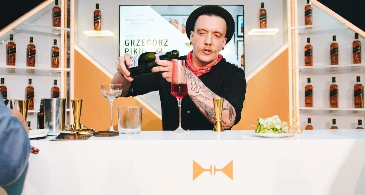 Barman Grzegorz Pikuła, w stylowym kapeluszu, nalewa drinka. W tle półki z butelkami whisky, idealne dla miłośników krwawych mary.