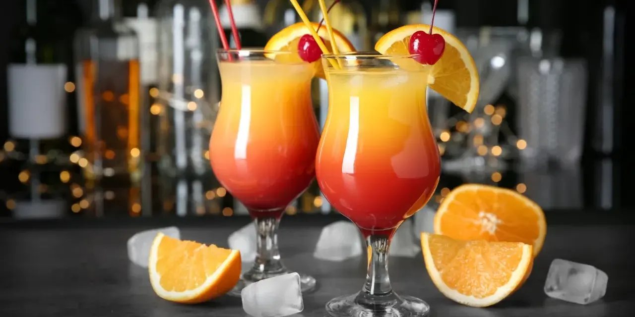Dwa kolorowe drinki z parasolką, ozdobione pomarańczą i wiśnią, stoją na barze.
