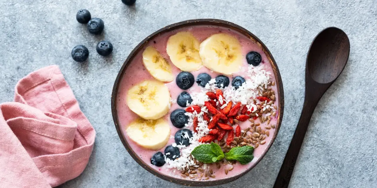 Kolorowy smoothie bowl z bananem, jagodami i kokosem, przypominający wyglądem tradycyjny kisiel. Obok leży drewniana łyżka i różowa serwetka.