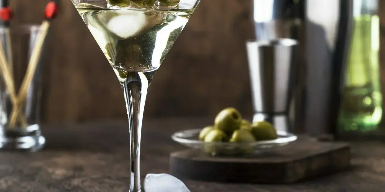 Elegancki drink z martini bianco, ozdobiony trzema oliwkami na patyczku. Obok kostki lodu i talerzyk z oliwkami.
