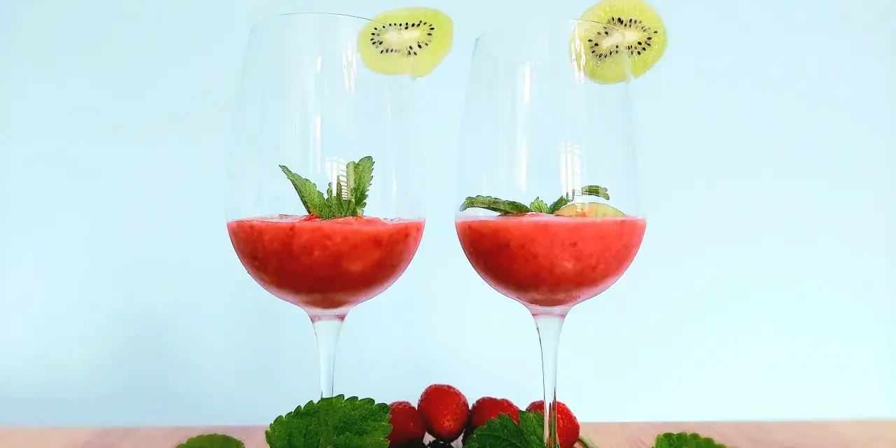Dwa kieliszki orzeźwiającego drinka truskawkowego z rumem, ozdobione plasterkami kiwi i miętą. Na drewnianym blacie leżą świeże truskawki i listki mięty.