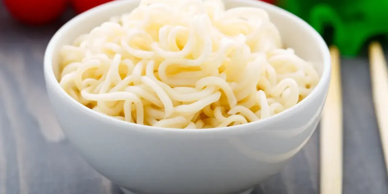 Biały makaron w misce, przypominający makaron konjac lub makaron z alg, z pomidorami i zielonymi warzywami w tle.