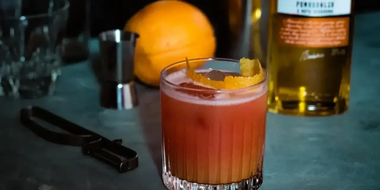 Orzeźwiający drink z likierem pomarańczowym, ozdobiony skórką cytrusową. Obok pomarańcza i butelka Saska.