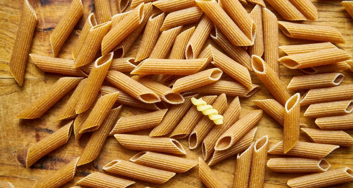 Makaron pełnoziarnisty vs zwykły. Na drewnianym tle leży mnóstwo makaronu penne. Wśród nich wyróżnia się jeden, żółty makaron fusilli.