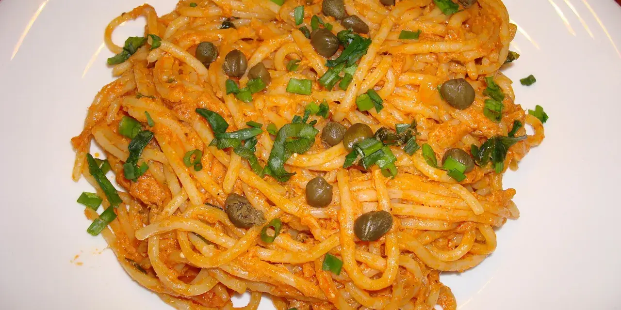 Pyszne spaghetti z tuńczykiem i kaparami, posypane szczypiorkiem, na białym talerzu.