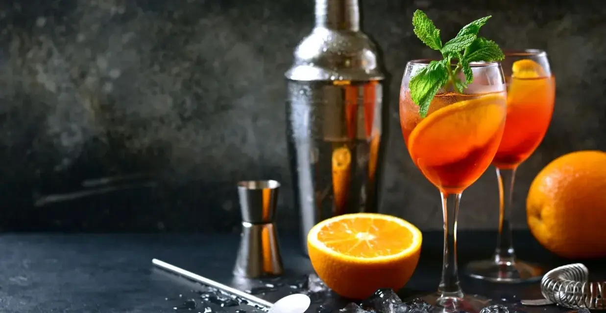 Orzeźwiający aperol z szampanem, udekorowany pomarańczą i miętą, gotowy do podania. Obok shaker i akcesoria barmańskie.