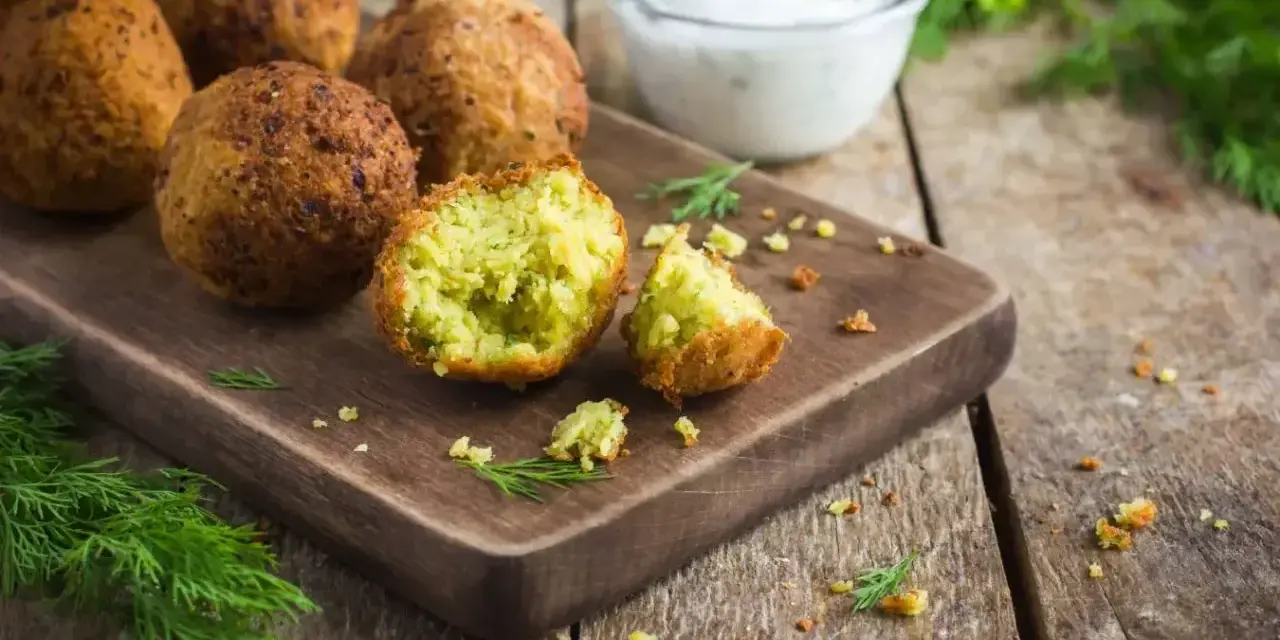 Chrupiące falafelki na desce, jeden przekrojony, ukazujący zielone wnętrze. Obok miseczka sosu i świeży koperek. Idealny falafel przepis.