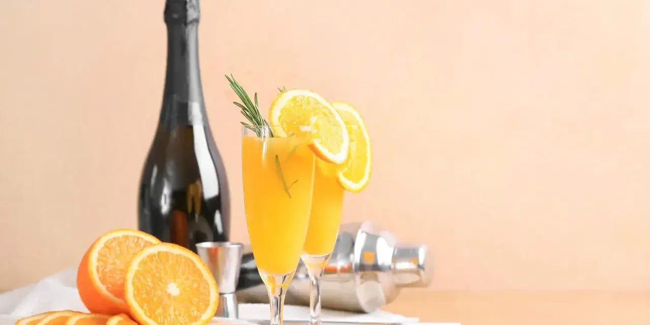 Dwa kieliszki z pomarańczowym napojem, ozdobione plasterkami pomarańczy i rozmarynem. Obok butelka szampana, shaker i świeże pomarańcze. Mimosa czy Bellini?