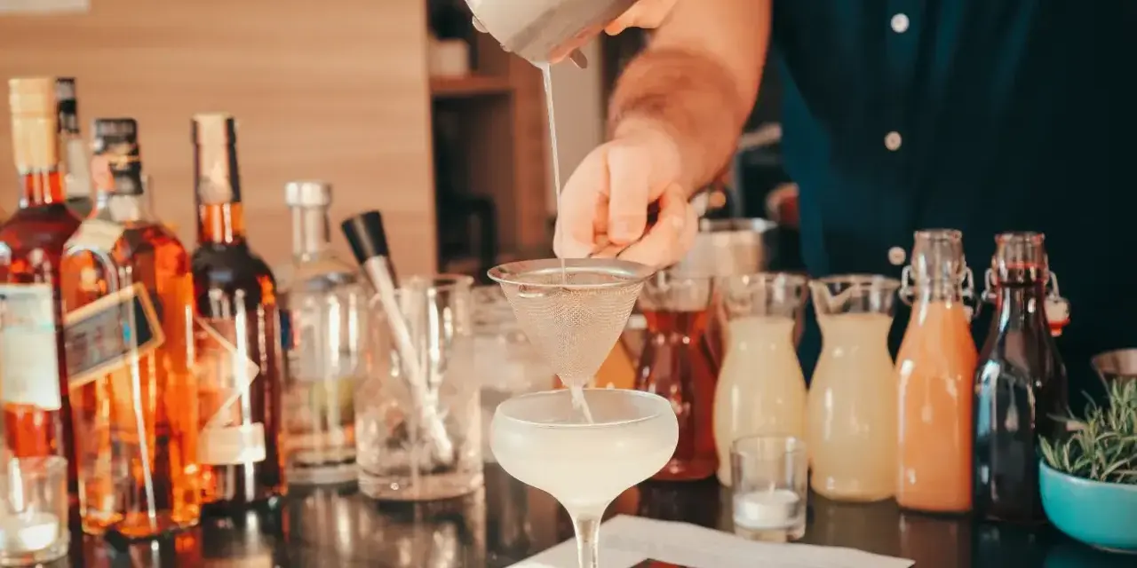 Barman przelewa płyn z shakera przez sitko do kieliszka, tworząc koktajl. W tle butelki alkoholi i kolorowe syropy.