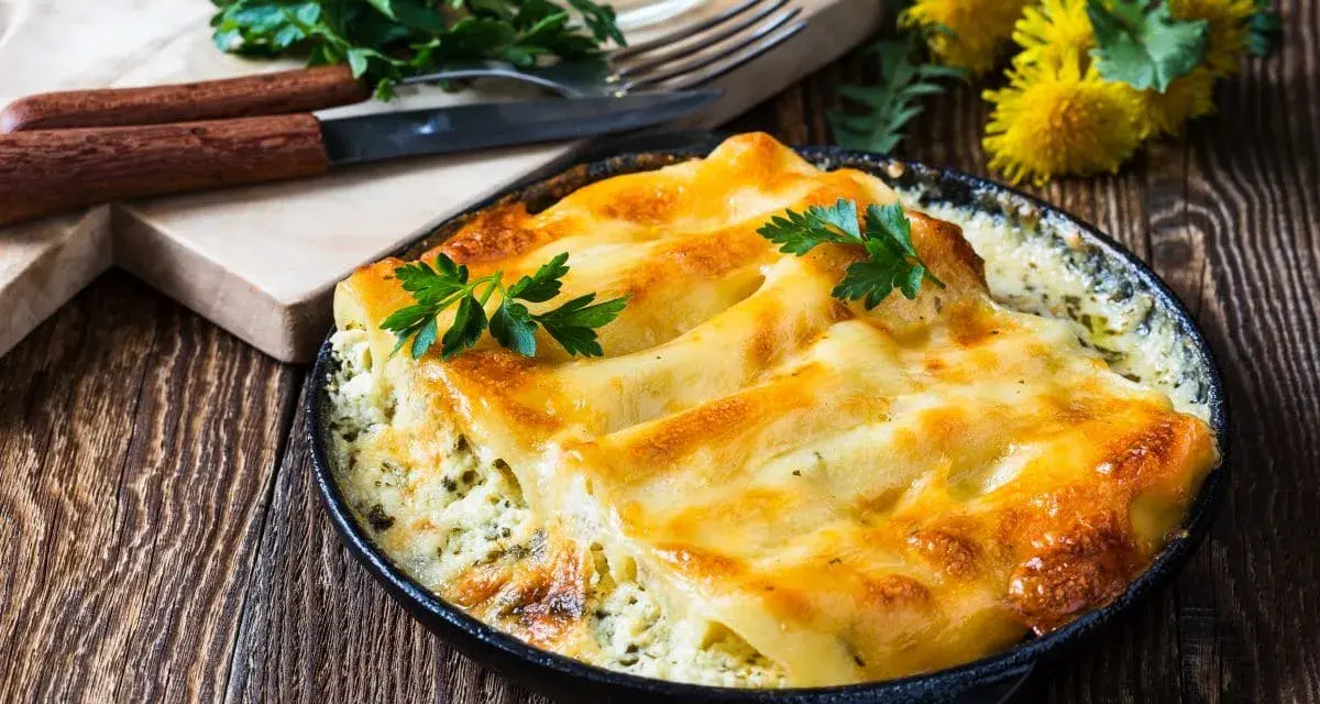 Pyszne conchiglioni zapieczone z serem i ziołami, podane w czarnej patelni. Obok deska z nożem i widelcem, oraz pęczek natki pietruszki.
