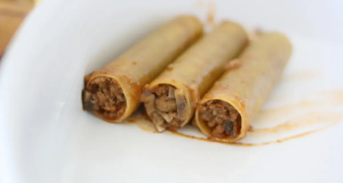 Trzy cannoloni z farszem mięsnym, wyglądające jak naleśniki z mięsem, leżą na białym talerzu.