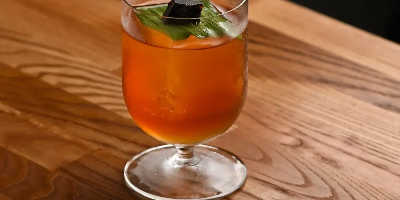Klasyczny Mai Tai z sokami, ozdobiony liściem i kostką ciemnego rumu, na drewnianym blacie.