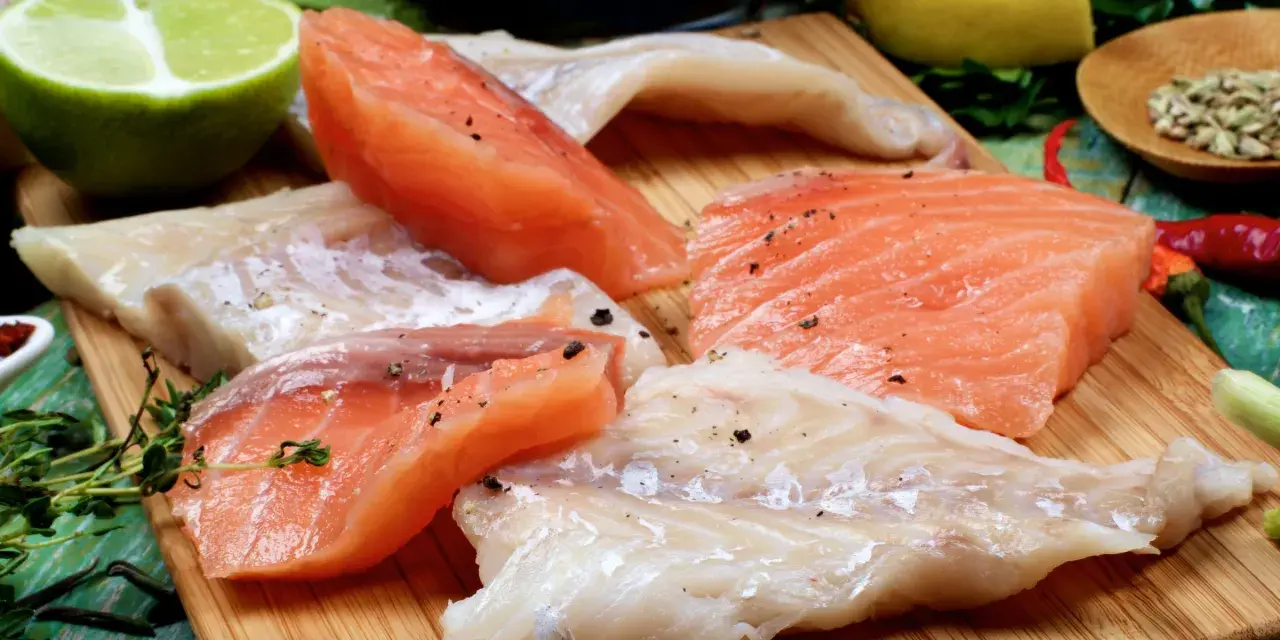 Stek z łososia kontra polędwica z dorsza. Surowe kawałki ryb, przyprawione pieprzem, leżą obok limonki, cytryny i ziół.