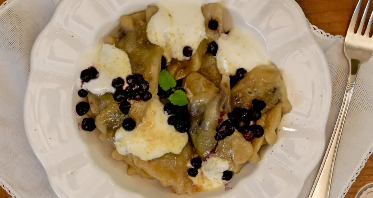 Knedle ze śliwkami czy pierogi z jagodami? Na talerzu pyszne pierogi z jagodami, polane śmietaną i posypane świeżymi owocami.