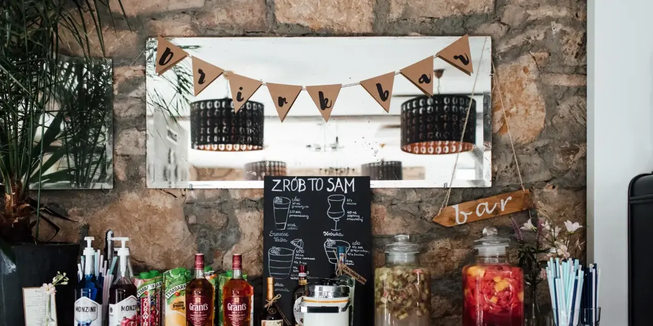 DIY drink bar z butelkami alkoholu, sokami i dzbankami z napojami. Tabliczka "ZRÓB TO SAM" zachęca do tworzenia własnych drinków.