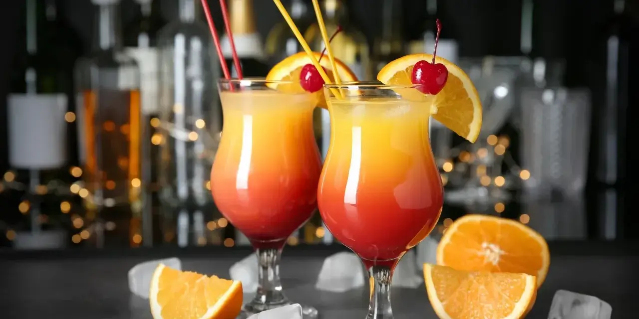 Dwa drinki z wódką, ozdobione pomarańczą i wiśnią, z kostkami lodu i owocami na blacie.