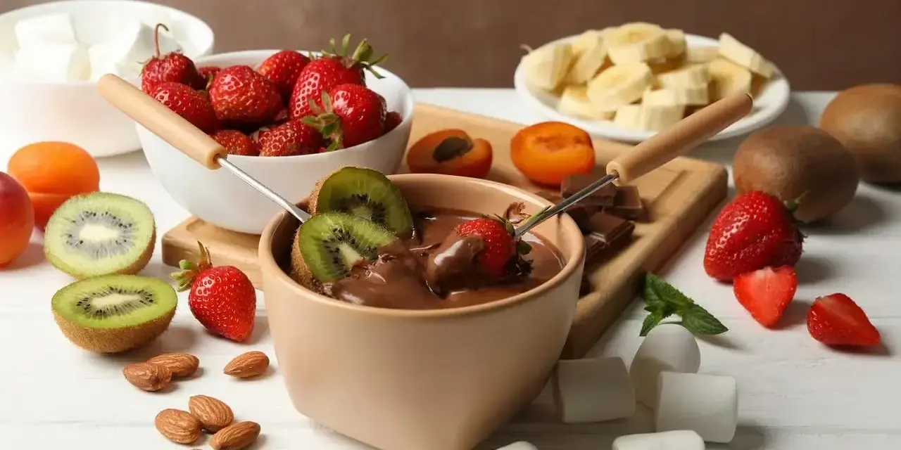 Jak zrobić fondue czekoladowe: zanurzaj truskawki i kiwi w gorącej czekoladzie. Na stole owoce, pianki i migdały.