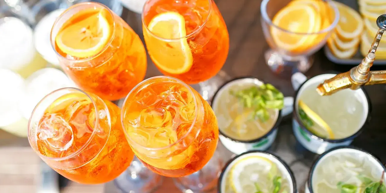 Orzeźwiający drink z prosecco, udekorowany plasterkiem pomarańczy, czeka na gości. Obok inne napoje z miętą i cytryną.