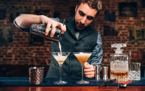 Barman przelewa drinka do kieliszka, pokazując, jak być dobrym barmanem. Na blacie stoją też inne drinki i akcesoria.