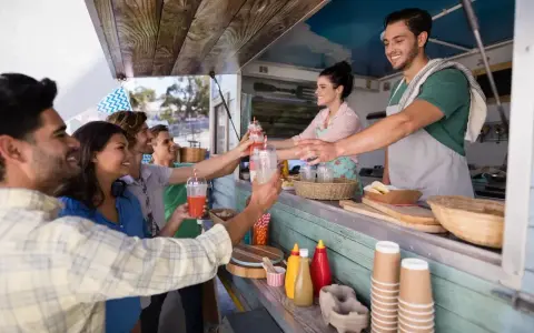 Uśmiechnięci klienci odbierają napoje od barmana w food trucku. Ciekawe, ile zarabia barman w takim miejscu?