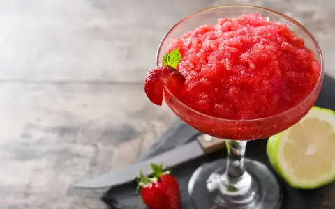 Orzeźwiający strawberry daiquiri, udekorowany plasterkiem truskawki i listkiem mięty, obok połówki limonki i świeżej truskawki.