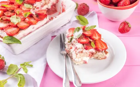 Tiramisu klasyczne kontra tiramisu truskawkowe. Porcja deseru z truskawkami i miętą na talerzu, obok naczynie z deserem i miseczka owoców.